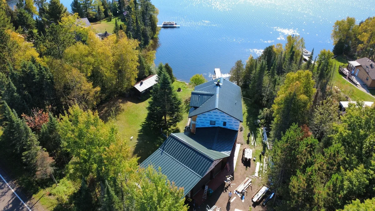 Aerial view of a blue standing seam metal roof on a wooded lakeside cabin in Brainerd, MN, with the lake visible beyond the treeline.