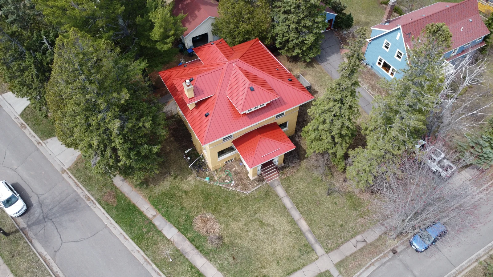 Tan brick two-story house topped with a vivid red standing seam metal roof seen from an angled aerial.