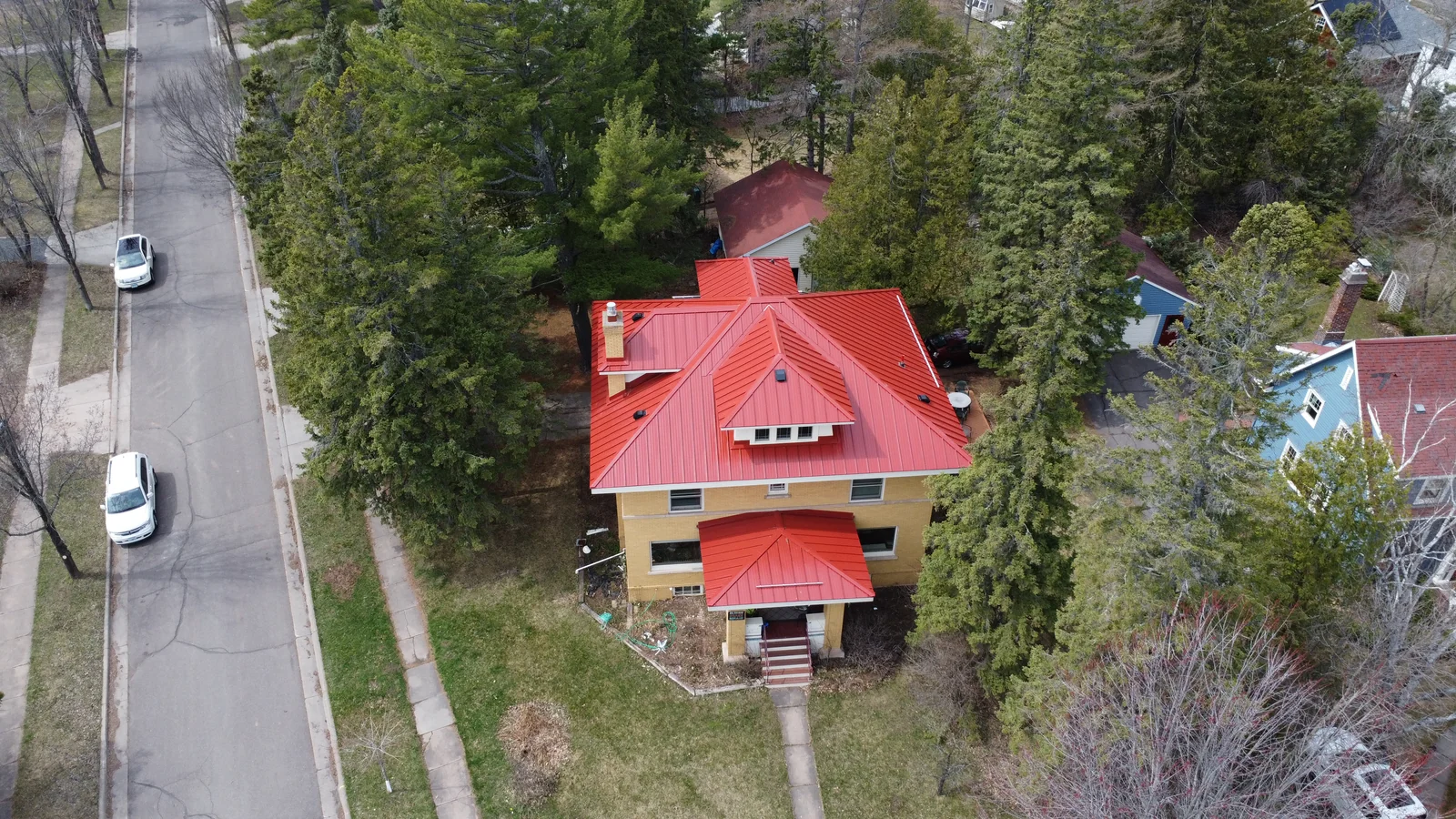 Red metal roof on the brick home viewed from above with the neighborhood street and pines alongside.
