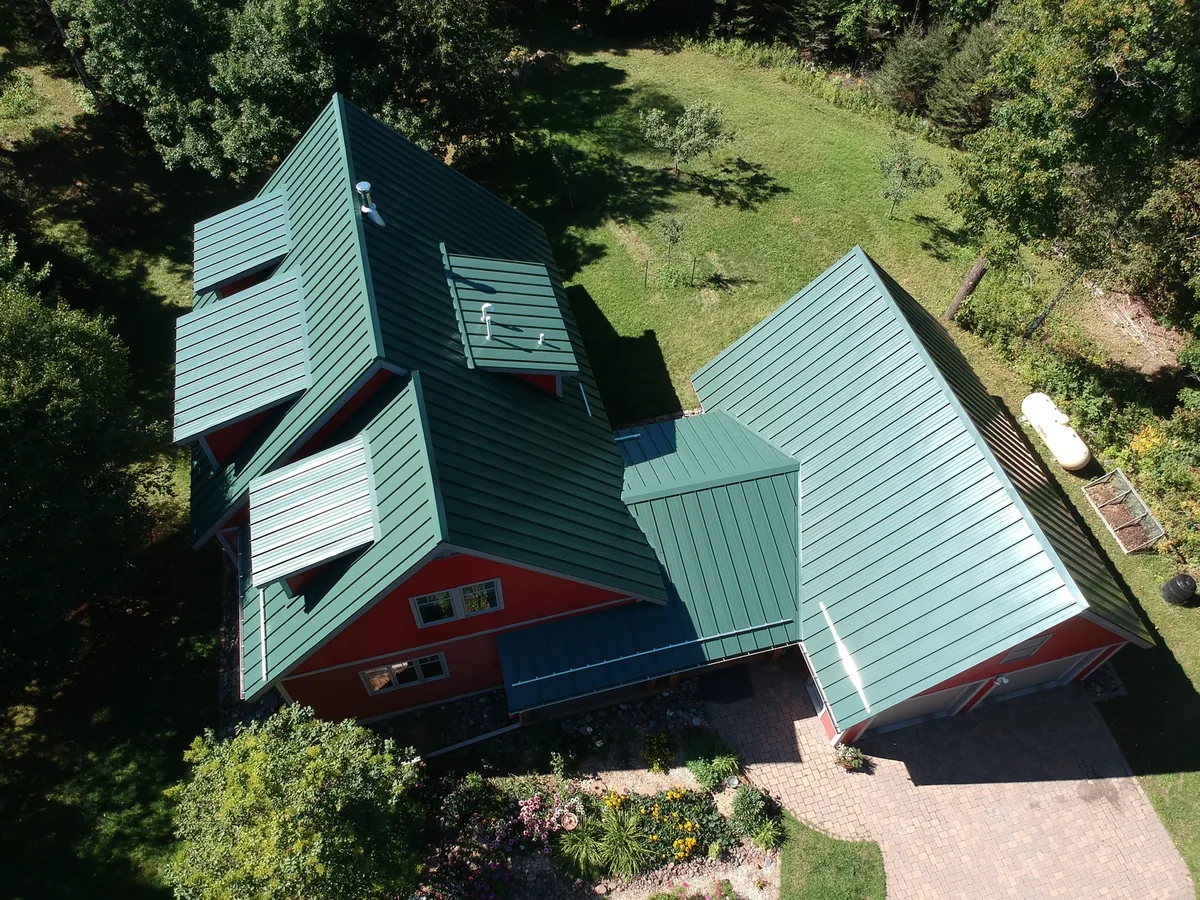 Steep gabled green standing seam panels meeting a lower hip section over the red house
