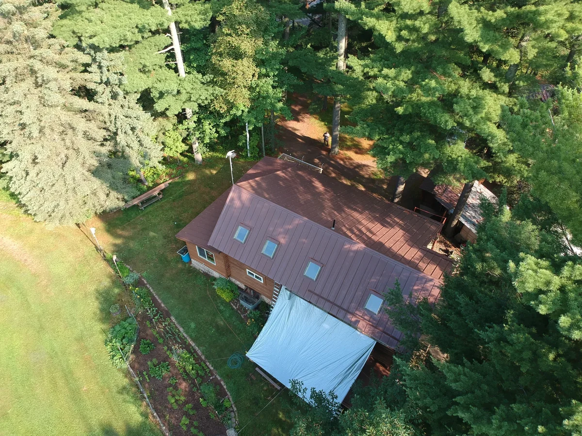 Angled aerial of the brown metal roof with a smaller outbuilding nearby in the trees