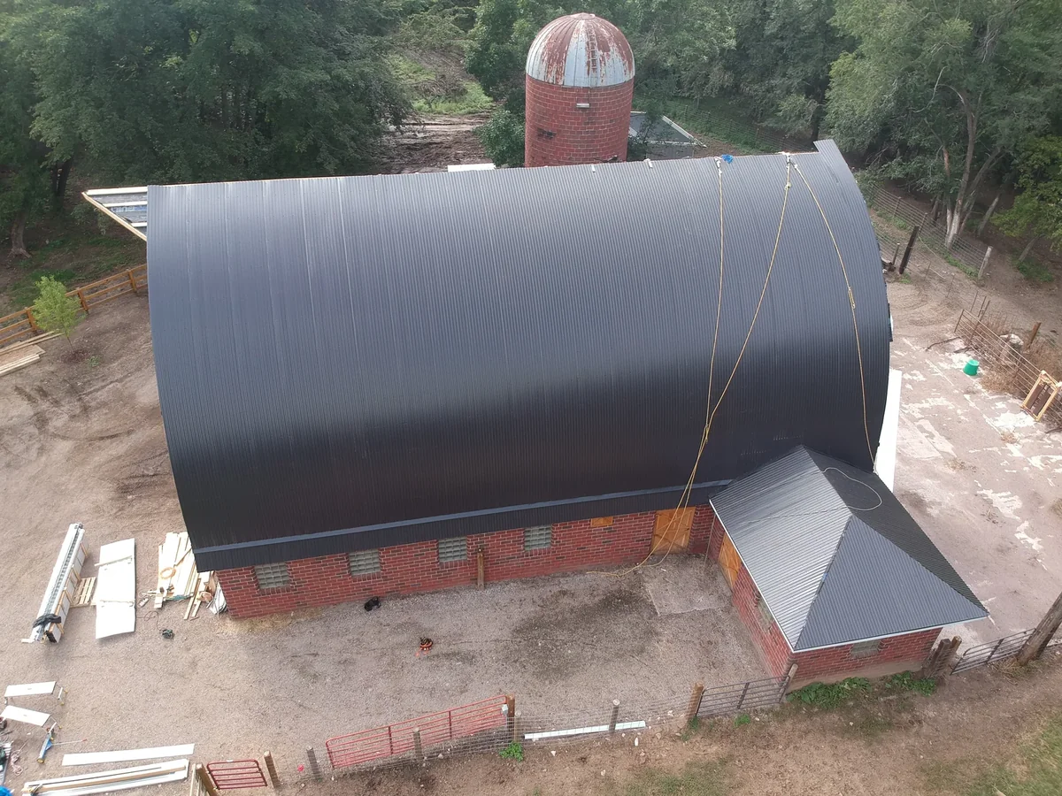 Aerial broadside of a historic arched barn with a new charcoal standing seam metal roof and brick base in Faribault, MN