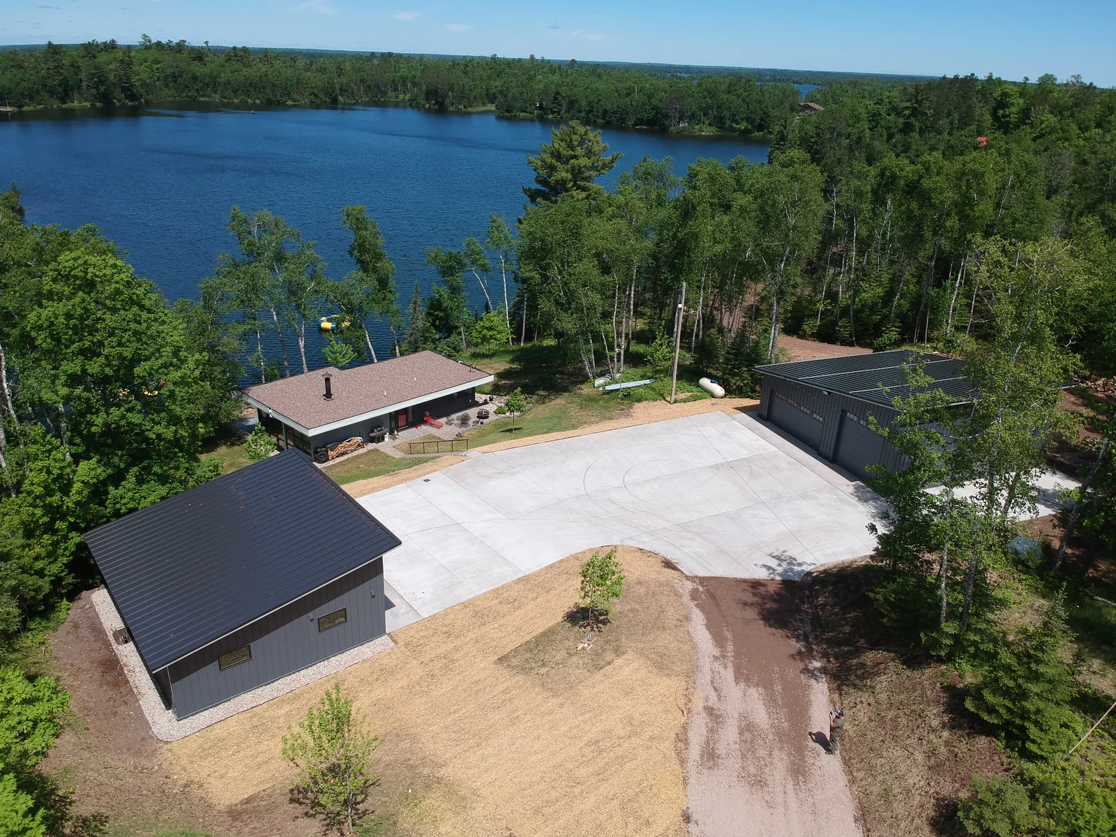 Aerial of the black-roofed building and its concrete drive with the lake glinting beyond the pines.