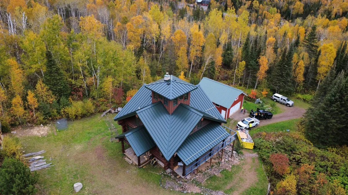 Drone shot of the log lodge and its grey standing seam metal roof set in the autumn woods