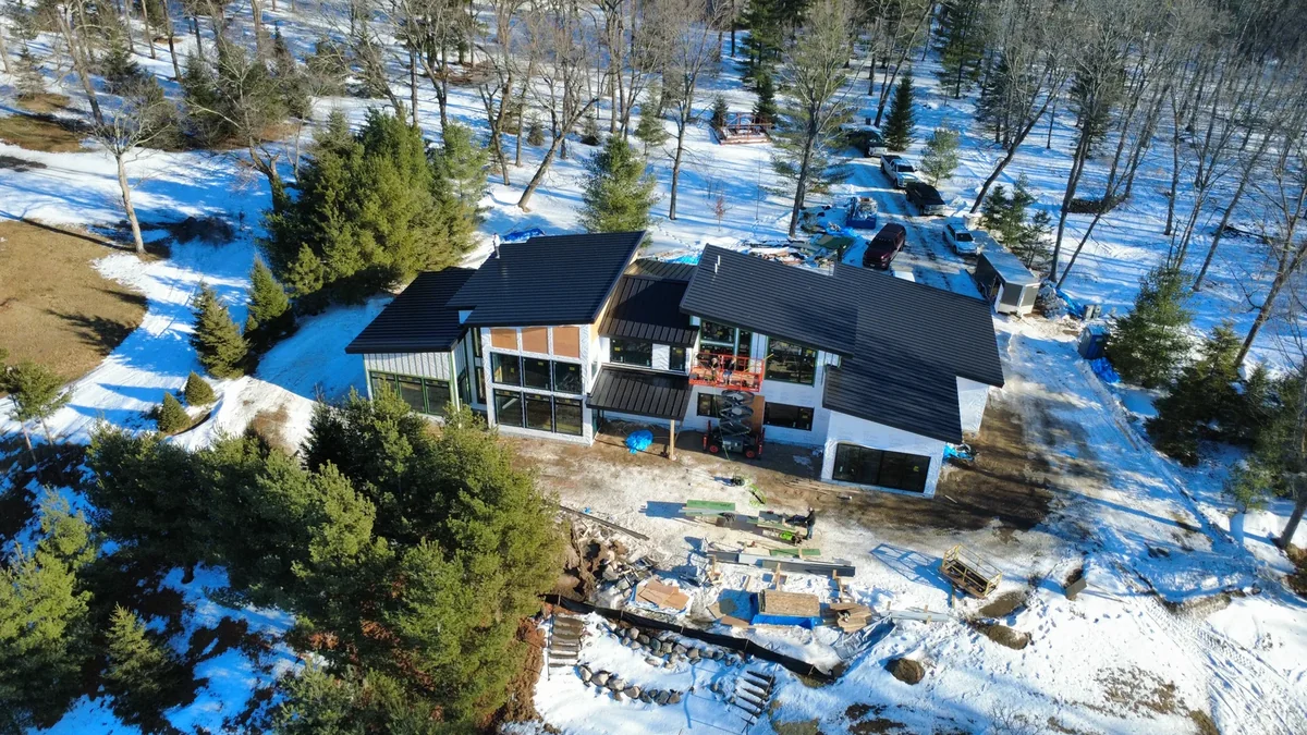 Aerial view of a dark metal roof on a residential shop under construction near Grand Rapids, MN, surrounded by patchy snow and pines.
