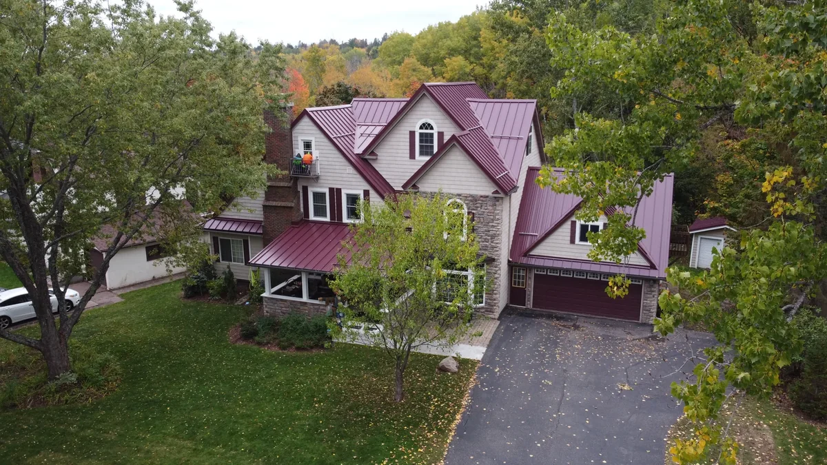 Aerial view of a two-story home with a deep red metal roof and wraparound porch in Hermantown, MN