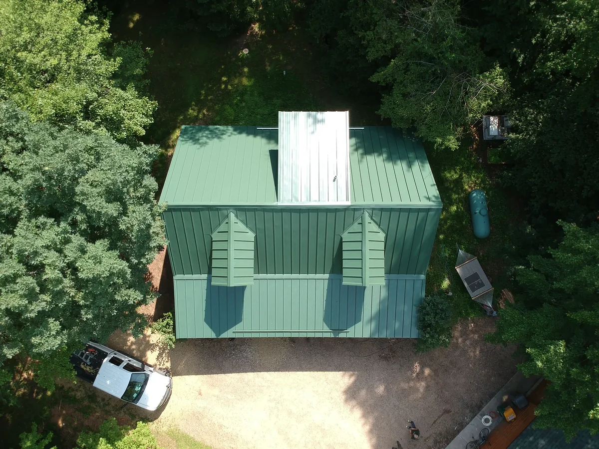 Straight-down view of the green metal roof and its white ridge feature surrounded by tall trees.