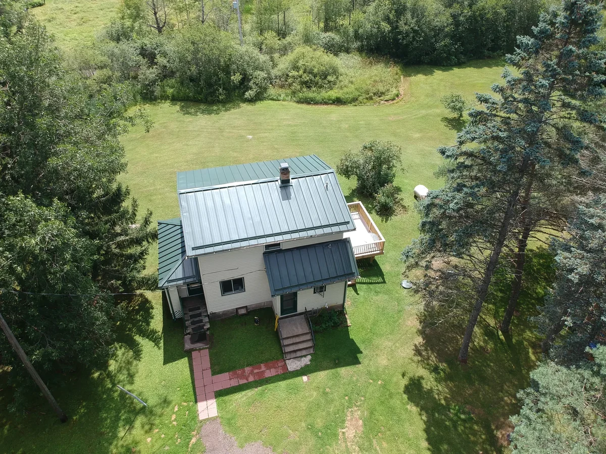 Aerial of a blue standing seam metal roof on a white two-story home amid a wooded yard in Lindstrom, MN.