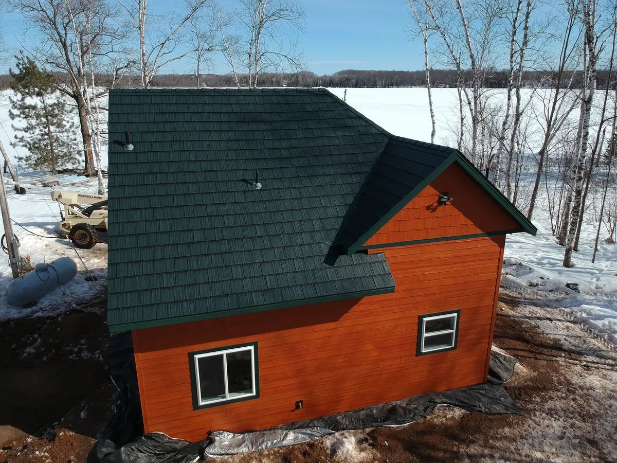 Aerial view of a cabin near Mora, MN with a dark green metal shingle roof over orange siding in winter