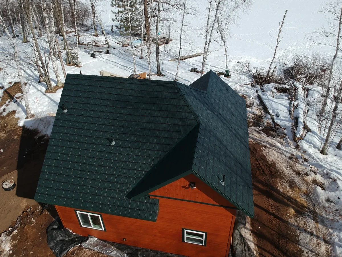 Drone shot of the green metal shingle roof on the orange cabin amid bare trees and snow