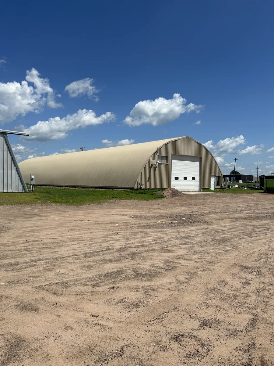 Wide front view of the rounded metal building with a white overhead door across a gravel yard