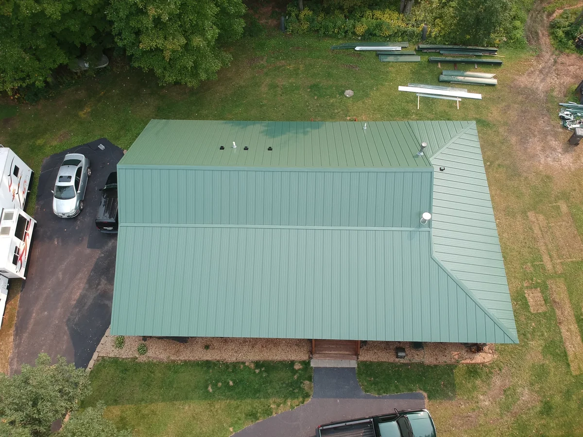 Aerial of a green standing seam metal roof on a wooded lot in Rice Lake, WI, with vehicles and materials nearby.