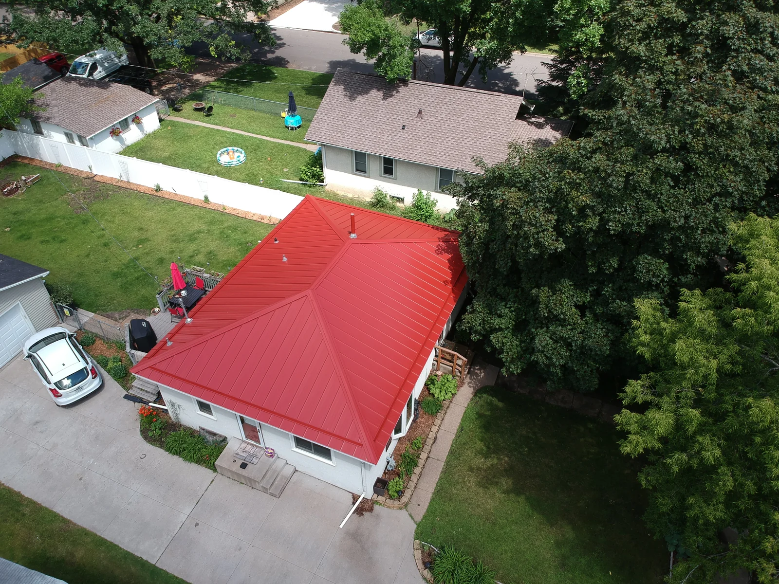 Drone shot of the red-roofed house with cars in the driveway and mature trees nearby.