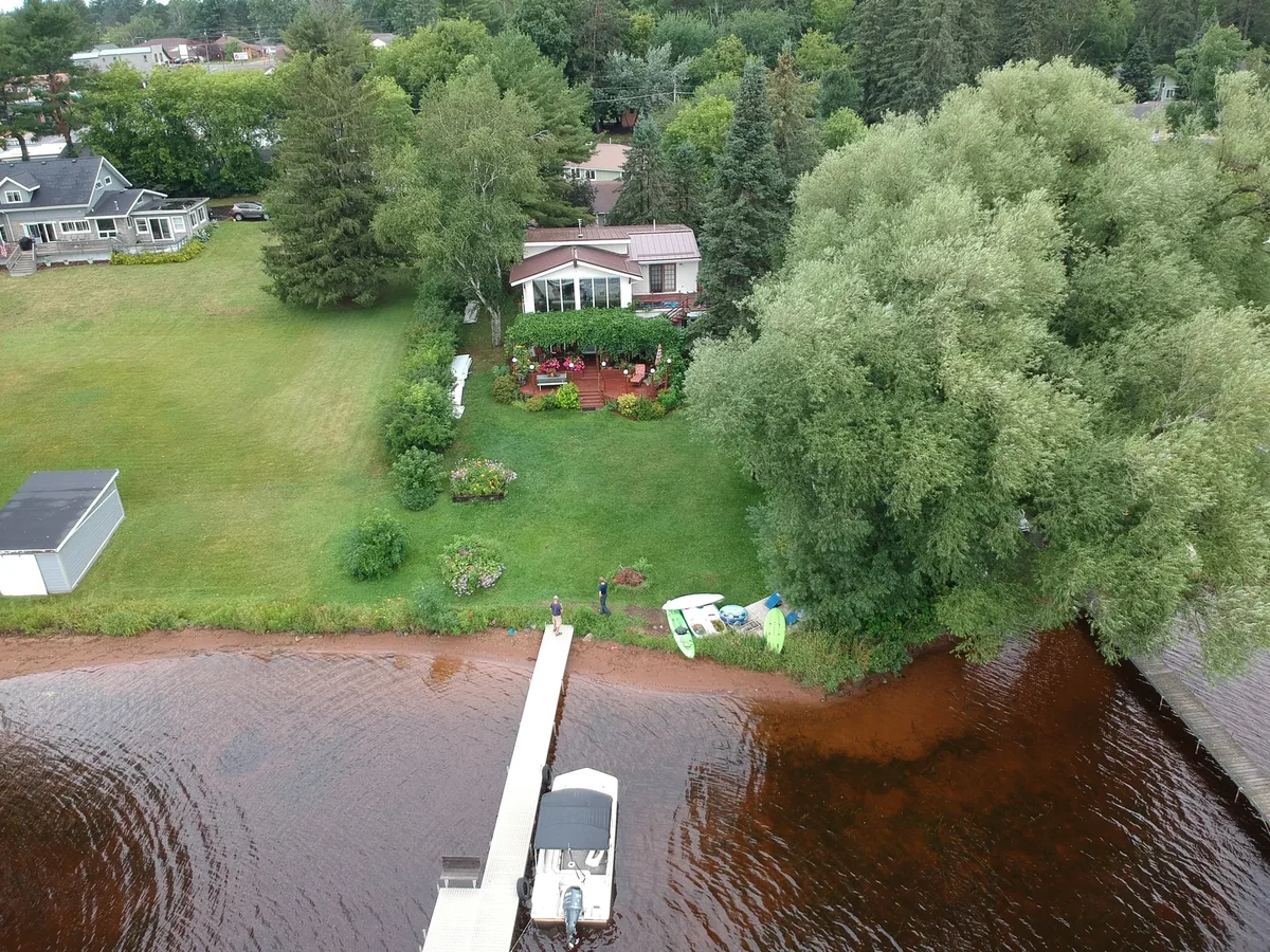 Wide drone shot of the property at the water's edge with the brown metal roof among the trees.