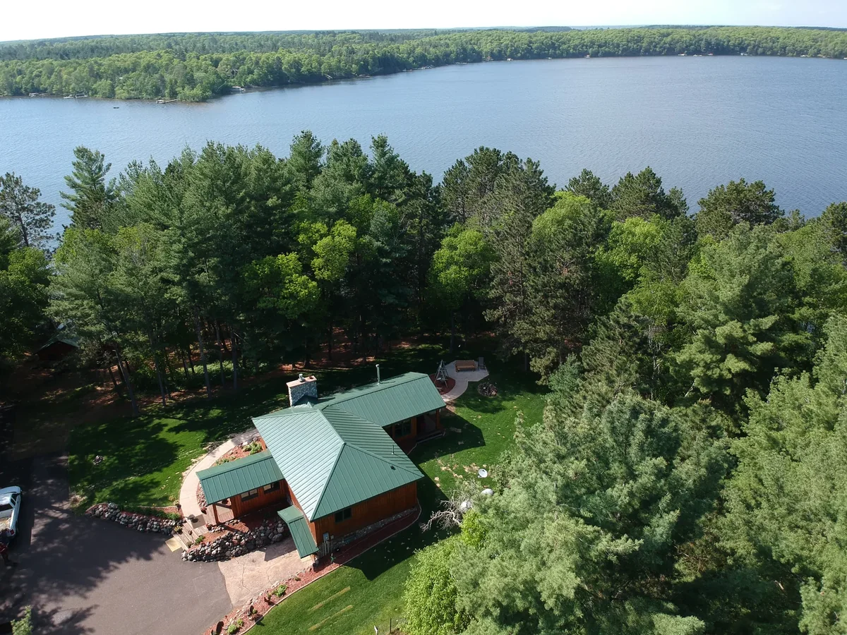 Aerial view of a green metal roof on a sprawling lakeside home near Turtle Lake, WI, with woods and water beyond.