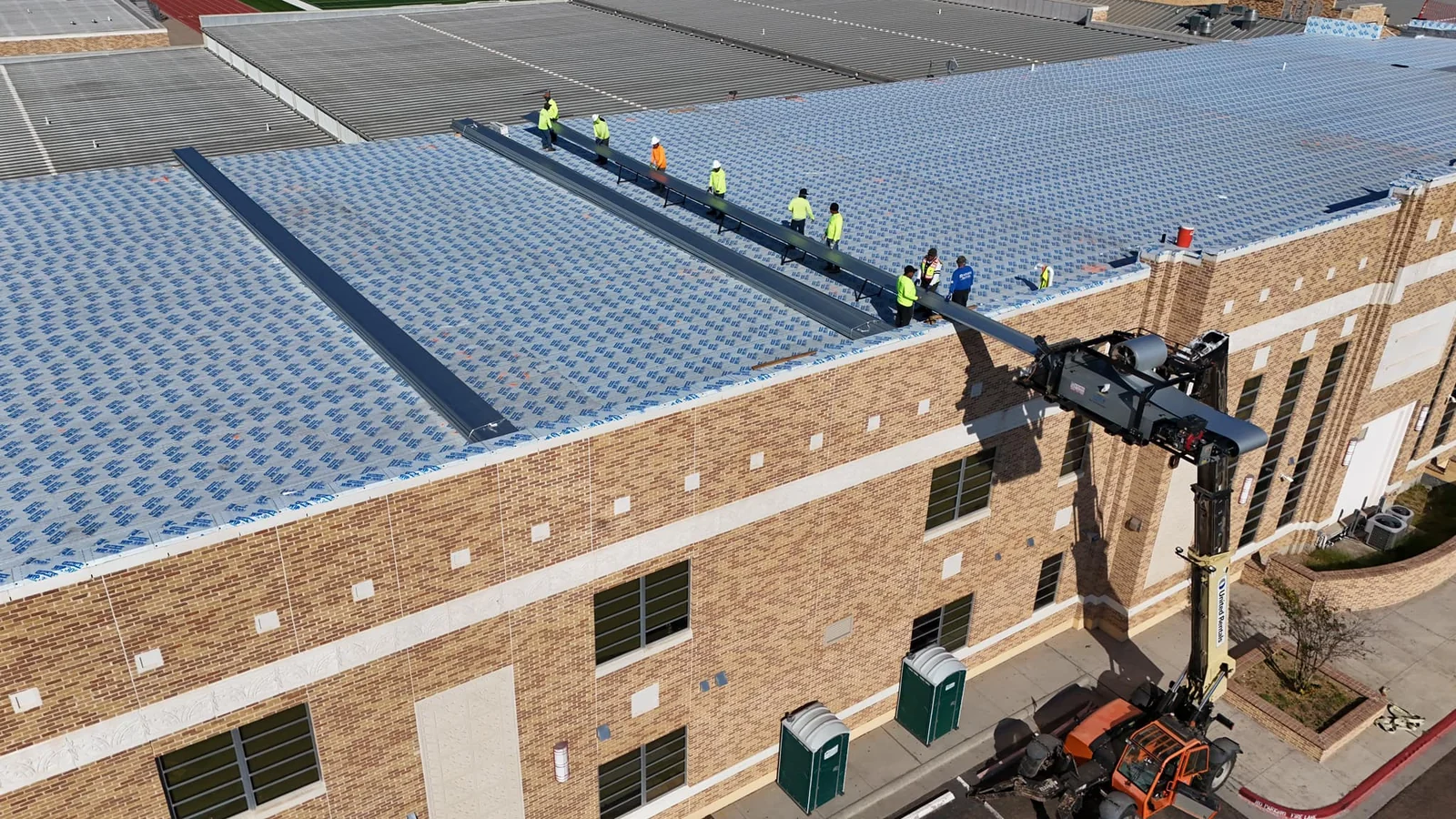 Roofing crew in safety vests installing metal panels as a crane lifts material to the rooftop.